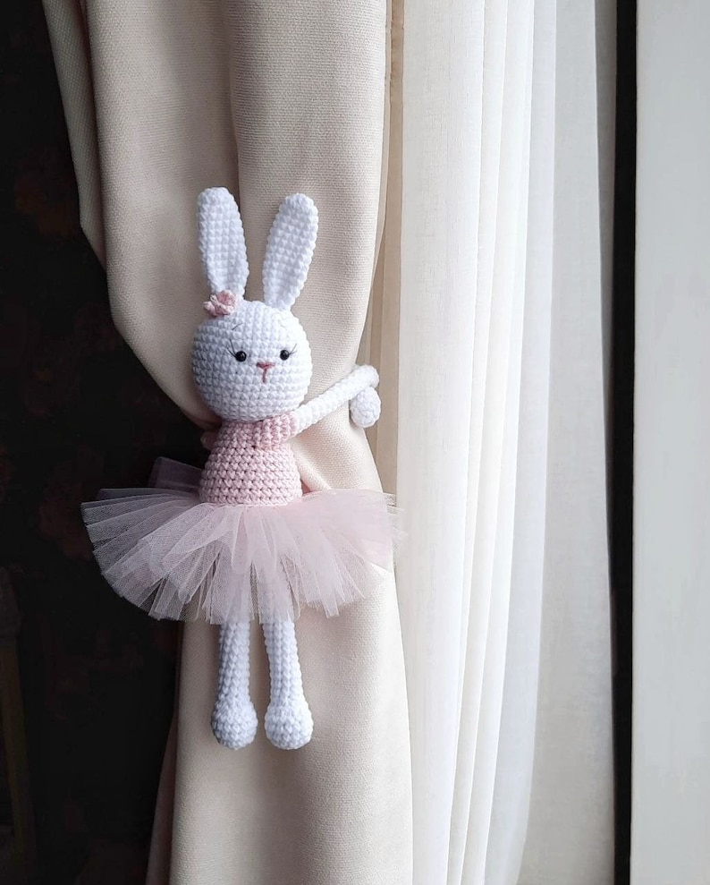 Puede incluir: Un conejito de crochet blanco con un tut&uacute; rosa y sujetando un amarrador de cortina. El conejito est&aacute; sujetando el amarrador con su brazo derecho y tiene una flor rosa en la cabeza.
