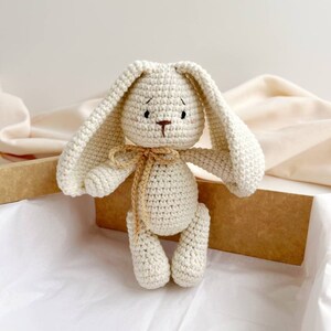 May include: A cream-coloured, crocheted bunny toy with long ears and a small, beige bow. The bunny has embroidered facial details and is presented in a brown box with white tissue paper.