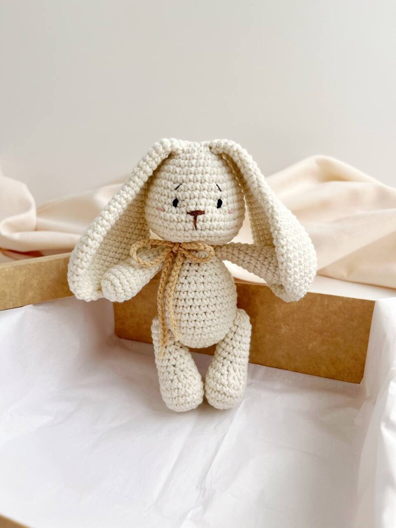 May include: A cream-coloured, crocheted bunny toy with long ears and a small, beige bow. The bunny has embroidered facial details and is presented in a brown box with white tissue paper.
