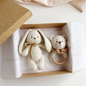 May include: A cream crochet bunny and bear rattle set in a brown cardboard box. The bunny has long ears and a bow, the bear rattle has a wooden ring. The box is lined with white paper, ideal for a baby gift.