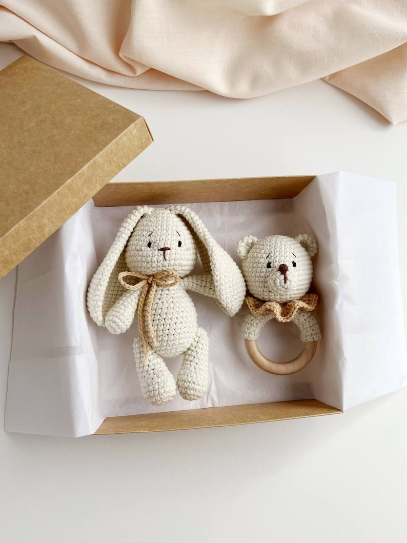 May include: A cream crochet bunny and bear rattle set in a brown cardboard box. The bunny has long ears and a bow, the bear rattle has a wooden ring. The box is lined with white paper, ideal for a baby gift.