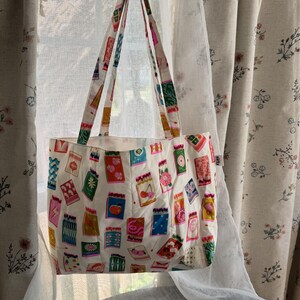 May include: A white fabric tote bag featuring a repeating pattern of colorful matchbox illustrations. The bag has two long shoulder straps and a small white rectangular tag on the side. The print includes various designs like cherries, apples, and geometric shapes.