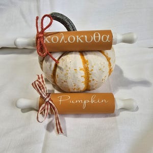 May include: Two decorative rolling pins with white handles and orange barrels, one labeled "Pumpkin" and the other with Greek text. They are adorned with red and brown ribbons and rest on a white and orange pumpkin.