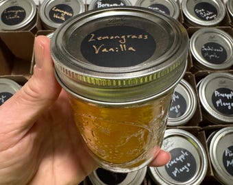 May include: A close-up of a glass jar filled with a yellow liquid. The jar has a silver lid with a black label that reads "Lemongrass Vanilla". The jar is surrounded by other jars with various labels.