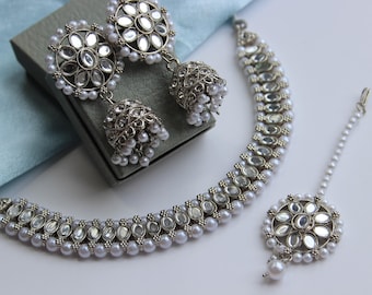 May include: A silver and white bridal jewelry set with a necklace, earrings, and a maang tikka. The necklace and earrings feature a floral design with pearl accents.