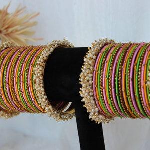 May include: A set of colourful bangles with a green, pink and orange striped pattern. The bangles are decorated with small gold beads and white pearl accents.