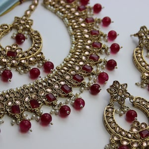 May include: A gold-tone necklace and earrings set featuring red and white stones. The necklace has a layered design with a central row of larger red stones. The earrings are dangle style with a similar design to the necklace.