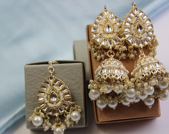 May include: Gold-toned jewelry set featuring a teardrop-shaped pendant and two pairs of earrings. The earrings have a bell shape and are adorned with white pearls and clear crystals. The set is displayed on a brown and gray box.