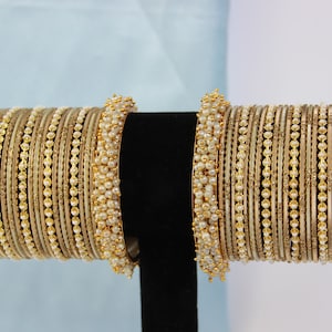 May include: A set of gold and white bangles with pearl accents. The bangles are stacked on a black stand.