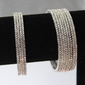 May include: A set of silver bangles with clear rhinestones. The bangles are stacked on a black display stand.