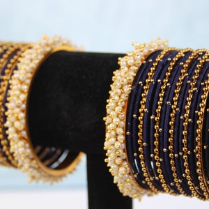 Può includere: Un set di braccialetti blu navy con dettagli dorati e perline bianche simili a perle. I braccialetti sono disposti in pile, mostrando il loro design intricato. I gioielli sono esposti su un supporto nero su sfondo azzurro.