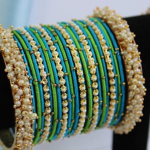 Könnte beinhalten: Ein Set aus grünen und blauen Seidenfaden-Armreifen mit goldenen Akzenten und weißen Perlenverzierungen.