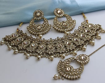May include: A gold and white Indian bridal jewellery set with a choker necklace, earrings, and a maang tikka. The set is adorned with sparkling stones and pearls.