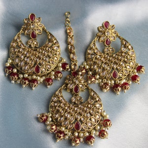Può includere: Un set di orecchini dorati e un maang tikka abbinato, con intricati disegni floreali e pietre rosse e bianche.