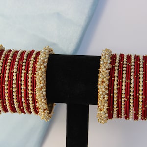 May include: Two sets of red bangles with white pearl accents. The bangles are displayed on a black stand against a white and light blue background. The bangles are a traditional style of jewellery.