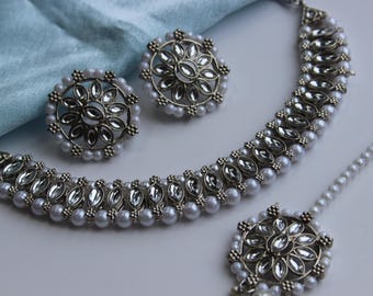 May include: A silver necklace, earrings, and a maang tikka set with white pearl accents and clear rhinestones. The jewelry is arranged on a light blue fabric background.