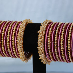 Op de afbeelding: Een set van marroon en roze zijden draad armbanden met gouden accenten en witte parels.