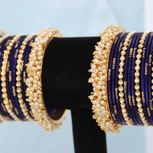 May include: Navy blue bangles with gold detailing and small, white pearl clusters. The bangles are presented on a black stand, highlighting their detailed design and elegant look. A stylish accessory.