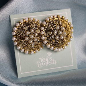 Peut inclure: Une paire de boucles d'oreilles de couleur or avec un motif circulaire. Les boucles d'oreilles sont ornées de perles blanches sur les bords et au centre. Les boucles d'oreilles sont présentées sur une carte bleu clair avec le texte "Tej's Trinkets".