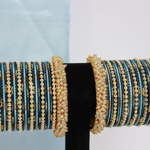 May include: A set of blue and gold bangles with white pearl accents. The bangles are stacked on a black display stand.