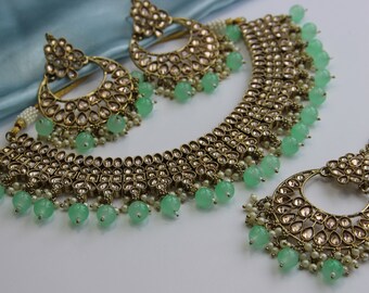 May include: A gold-tone necklace, earrings, and a maang tikka set with green and white beads. The necklace has a layered design with multiple rows of stones and beads.