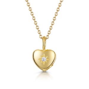 May include: A gold heart pendant necklace. The heart-shaped pendant has a starburst design with a small, clear gemstone in the middle. The necklace chain is gold and is attached to the pendant.