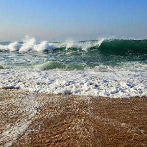 Può includere: Vista panoramica di una spiaggia con onde che si infrangono. L'acqua è un mix di turchese e verde scuro, con schiuma bianca. La sabbia è marrone chiaro e il cielo è azzurro.