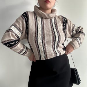 Può includere: Un maglione beige a collo alto con righe verticali marroni, nere e bianche. Il maglione ha un collo alto e polsini a coste, con accenti fantasia sulle maniche. La persona indossa una gonna nera e una borsa nera.
