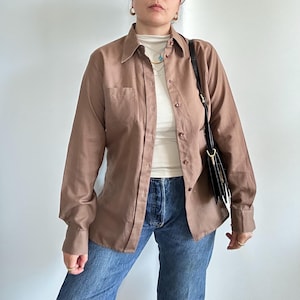 Può includere: Una camicia marrone con bottoni e taschino sul petto. La camicia è indossata sopra un dolcevita bianco e abbinata a jeans blu. Una borsa a tracolla nera è appesa di lato. La camicia ha maniche lunghe e un colletto classico.