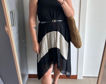 May include: A black and white pleated dress with a black belt. The dress has a high neckline and a high-low hemline. The model is wearing black sandals.