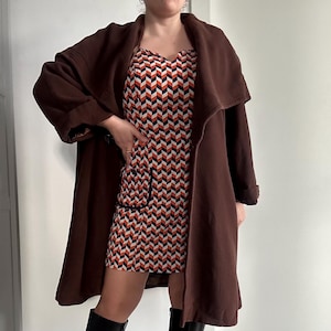 Può includere: Un cappotto marrone a maniche lunghe con un ampio colletto e un vestito con motivo geometrico sotto.