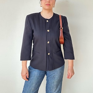 Può includere: Un blazer blu navy scuro con maniche a tre quarti e bottoni dorati. Il blazer ha un colletto rotondo e due piccole tasche. Una borsa in pelle marrone è appesa alla spalla.