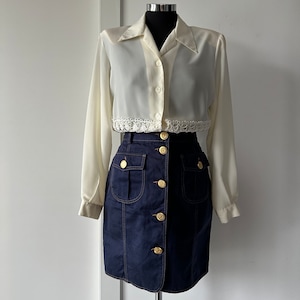 Può includere: Una blusa bianca con bottoni e un bordo in pizzo e una gonna in denim blu scuro con bottoni dorati.