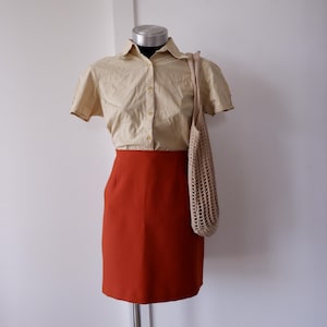 Può includere: Un manichino espone una camicia a maniche corte a quadri beige e bianca, abbinata a una gonna a tubino color ruggine. Una borsa tote beige a trama aperta è appesa alla spalla. L'outfit è presentato su sfondo bianco.