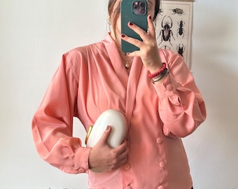 May include: A pink satin blouse with long sleeves and a double breasted closure. The blouse has a relaxed fit and is styled with a white clutch.