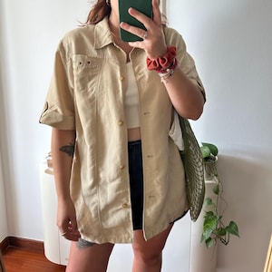 Può includere: Una camicia utility beige con bottoni e maniche corte. La camicia ha una tasca sul petto sinistro e una chiusura con bottoni dorati.