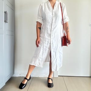 Puede incluir: Un vestido camisero blanco con mangas enrolladas. El vestido, a la altura de la rodilla, tiene un corte relajado. Un pequeño bolso rojo cuelga de un hombro. Los zapatos Mary Jane negros completan el look. El vestido es de tejido ligero.