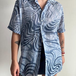 Può includere: Una camicia abbottonata di colore azzurro chiaro semitrasparente con un motivo astratto a spirale. La camicia ha maniche corte e un colletto.
