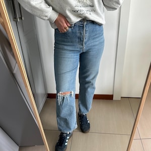May include: Light blue denim jeans with a distressed knee, paired with navy blue sneakers with white stripes. A gray sweatshirt with a graphic print is visible. The image is reflected in a wooden-framed mirror.