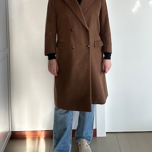 Può includere: Un lungo cappotto marrone doppiopetto con un dolcevita nero sotto. La persona indossa jeans blu e scarpe beige.