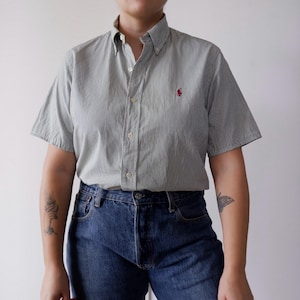 Può includere: Camicia a maniche corte con motivo a righe verticali grigie e bianche. La camicia presenta un piccolo logo rosso sul petto. La modella indossa jeans blu.