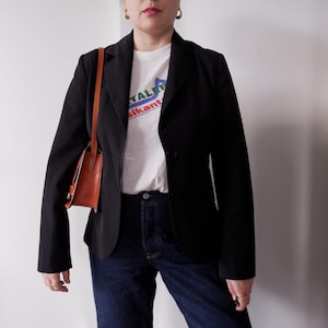 May include: A black blazer with a two-button closure, paired with a white graphic t-shirt and dark blue jeans. A small, brown leather purse with a shoulder strap is also visible.