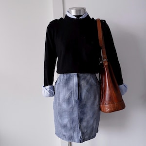 Può includere: Un maglione nero sopra una camicia azzurra, abbinato a una gonna a quadretti blu e bianca. Una borsa a tracolla in pelle marrone completa l'outfit, mostrando uno stile classico e preppy.
