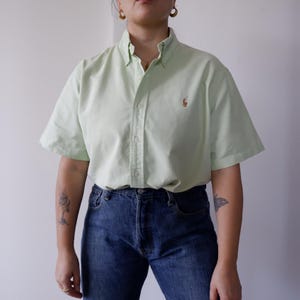 Può includere: Una camicia verde chiaro a maniche corte con un piccolo logo sul petto, abbinata a jeans blu. La camicia ha un colletto button-down e una vestibilità rilassata. I jeans sono un classico modello a cinque tasche.