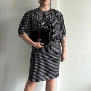 Op de afbeelding: Een zwart-witte polkadotjurk met een bijpassende top en een zwarte handtas. De handtas heeft een rood-groene streep en een goudkleurige sluiting. De jurk heeft korte mouwen en een knielange rok.