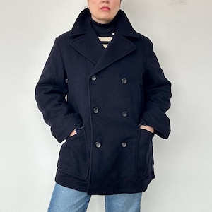 Puede incluir: Un abrigo de paño azul marino de doble botonadura con solapas grandes y dos filas de botones oscuros. El abrigo tiene dos bolsillos delanteros y un diseño clásico y entallado. El modelo lleva un cuello alto negro y vaqueros azules.