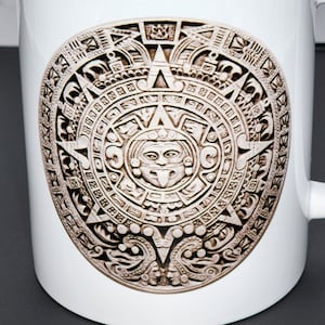 May include: White ceramic mug featuring a detailed, beige-colored Aztec calendar design. The mug has a curved handle and a cylindrical shape. The Aztec calendar design is centered on the mug's surface.
