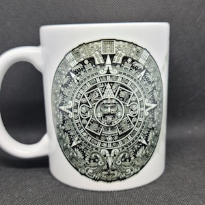May include: White ceramic mug featuring a detailed black and gray illustration of the Aztec calendar. The design is centered on the mug, with a curved handle on the left side. The mug is set against a dark gray background.