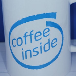 Puede incluir: Taza de café de cerámica blanca con un diseño azul que dice "coffee inside". El diseño está dentro de un óvalo azul. La taza tiene un asa y está sobre un fondo azul.