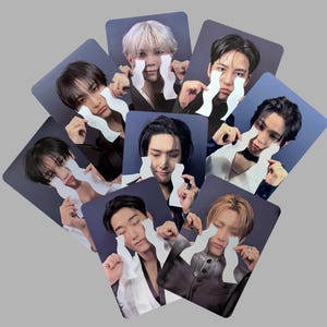 以下が含まれることがあります： 様々な人物の肖像画が描かれた正方形のカードのコレクション。各カードには、白い波型の紙の切り抜きが目に当てられています。カードは濃い青色の背景で、円形に配置されています。